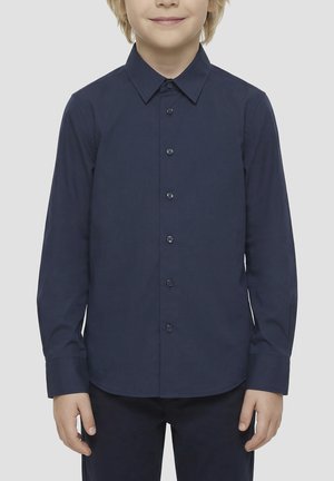 Enfant portant une chemise bleu marine à manches longues boutonnée, debout devant un fond clair uni, souriant légèrement.