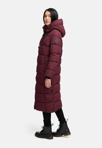 Cappotto piumino bordeaux con cappuccio, presenta sezioni trapuntate e cordini regolabili. Abbinato a pantaloni neri e stivali alla caviglia con lacci.