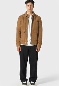 Brązowa zamszowa kurtka z kołnierzem, z przodu na zamek i dwiema kieszeniami na piersi. Nosi się ją na białej koszulce, w połączeniu z czarnymi spodniami o szerokich nogawkach i trampkami.