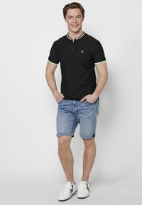 Juodas polo marškinėlis su žaliais akcentais, trumpomis rankovėmis ir mygtukais, derinamas su šviesiai mėlynomis džinsinėmis šortais ir baltais sportiniais bateliais.