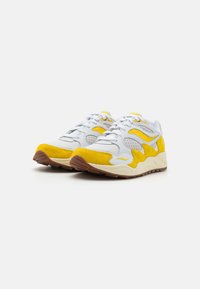 Sportschuhe mit einem überwiegend weißen Obermaterial, gelben Wildlederakzenten, strukturiertem Netzstoff und einer klobigen braunen Gummisohle.