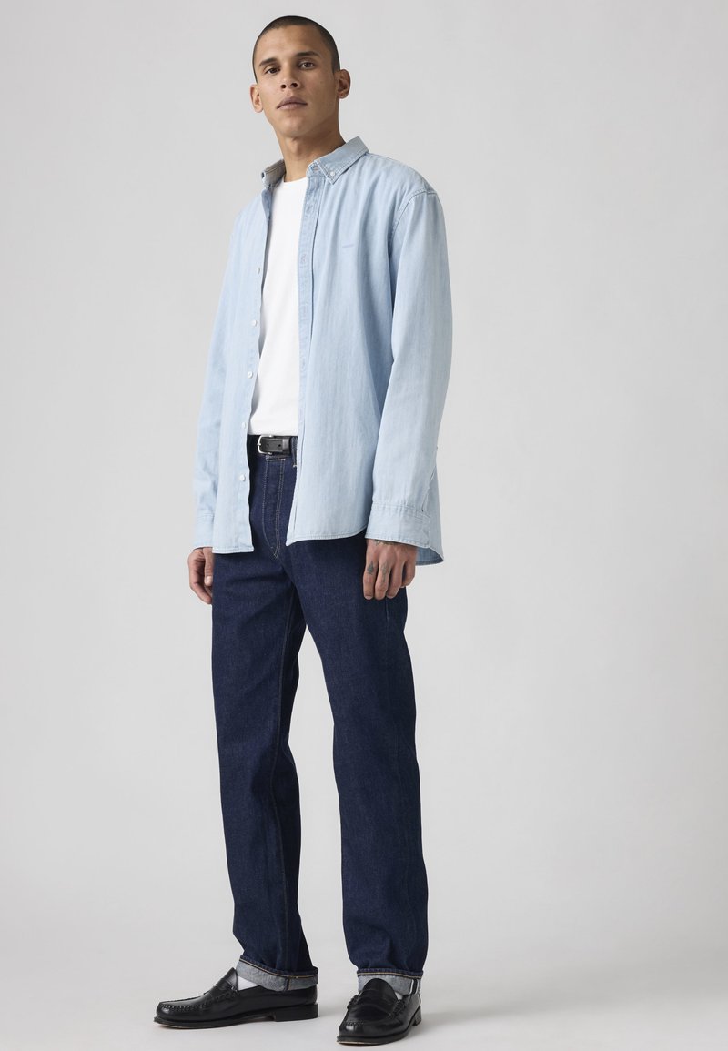 Hombre con camisa azul claro abotonada sobre camiseta blanca, vaqueros oscuros con puños, cinturón negro y mocasines negros, de pie frente a un fondo gris.