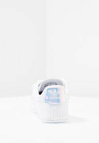 Vita Adidas-sneakers sedd från baksidan med en iridiserande hälflik som visar Adidas-logotypen och texturerad sula på en vit yta.