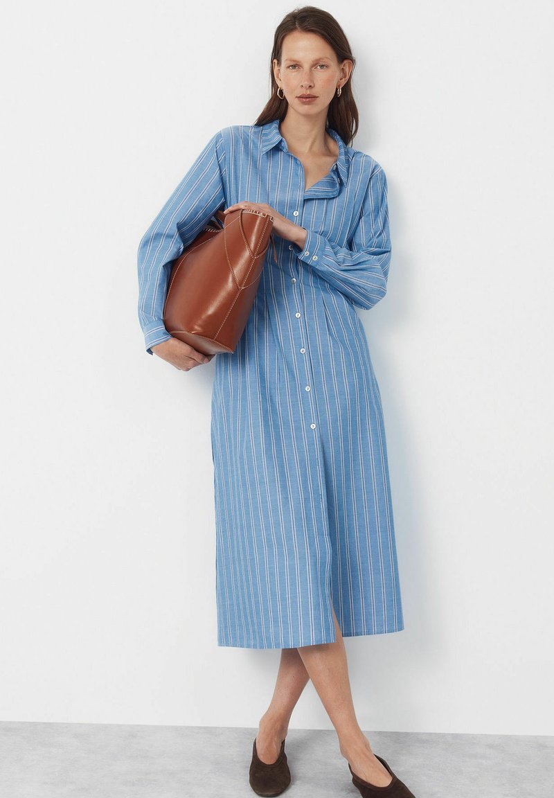 Vrouw met een blauw gestreepte lange overhemdjurk, die een grote bruine leren handtas vasthoudt, staand tegen een witte muur.