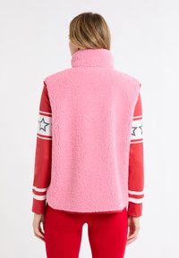 Różowa kamizelka z polaru z wysokim kołnierzem; miękka teksturowana tkanina; noszona na czerwonej bluzce z długim rękawem z białymi i czarnymi gwiazdkami.