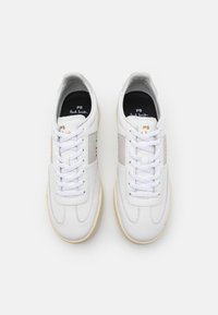 Par de zapatillas de piel blancas con cordones blancos, suela beige, paneles laterales grises, rayas multicolores y texto dorado "PS Paul Smith" en la lengüeta y en el lateral.