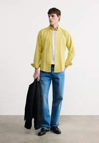 Νέος άνδρας με καστανά μαλλιά στέκεται φορώντας ένα κίτρινο πουκάμισο με κουμπιά πάνω από ένα λευκό μπλουζάκι, μπλε τζιν, μαύρα παπούτσια, κρατώντας ένα μαύρο μπουφάν στο ένα χέρι.