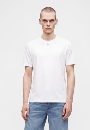 Joven con el pelo corto, vestido con una camiseta blanca lisa y vaqueros azules, de pie frente a un fondo claro.