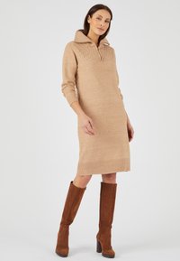 Abito a maglia color cammello con colletto alto e orlo a coste. Maniche lunghe e parte superiore testurizzata; abbinato a stivali marroni al ginocchio.