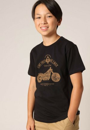 Tienerjongen met zwart haar draagt een zwart T-shirt met een bruine motorfietsafbeelding en de tekst "Live Fast Die Last" en beige broek.