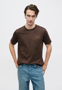 Καφέ βαμβακερό t-shirt με στρογγυλή λαιμόκοψη, κοντά μανίκια και μικρό πορτοκαλί τυπωμένο λογότυπο στο στήθος. Συνδυασμένο με ανοιχτόχρωμο μπλε τζιν.