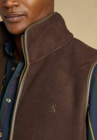 Hnedá fleece vesta s vysokým golierom, zapínaním na zips a kontrastným textúrovaným lemovaním. Má vyšité logo na prednej strane.