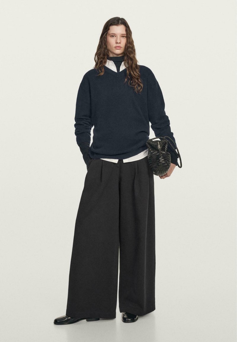 Marineblauwe trui met V-hals, gecombineerd met een witte blouse en zwarte wijde broek. Bevat een gestructureerde zwarte handtas en glanzende schoenen.