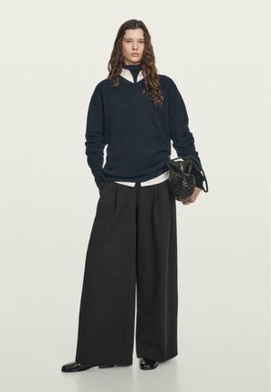 Maglione blu navy con scollo a V, abbinato a una camicia bianca con colletto e pantaloni neri a gamba larga. Caratterizzato da una borsa nera testurizzata e scarpe lucide.