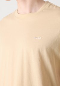 Beige tričko s kulatým výstřihem a malým bílým logem "HUGO" na levé straně hrudi, které nosí osoba.