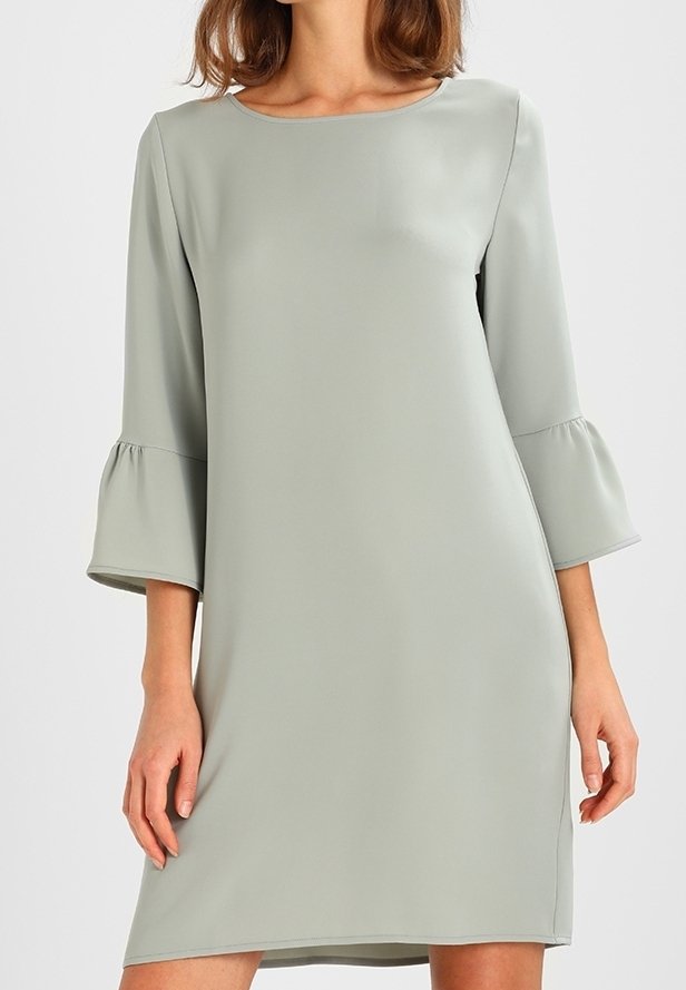 Femme portant une robe gris clair longueur genou avec des manches cloche trois-quarts et un col rond sur un fond uni.