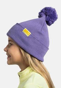 Purple knit beanie with a pom-pom on top and a yellow rectangular label featuring a mountain graphic. Soft texture, snug fit.