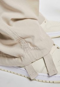 Sneaker beige con pelle testurizzata e suola bianca. Pantaloni in tessuto beige larghi scendono sopra la scarpa, evidenziando i dettagli di design.