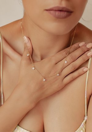 Goldene Kettennacklace mit fünf kleinen Blumenanhängern mit weißen Akzenten. Die Hand des Models verdeckt teilweise die Halskette und zeigt ihr zartes Design.