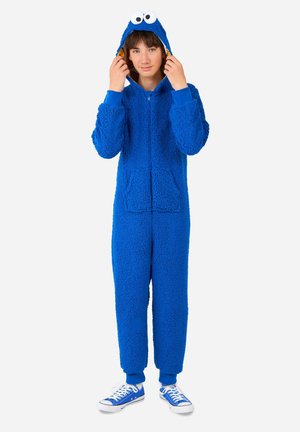Blauwe fleece jumpsuit met een gestructureerd oppervlak, capuchon met grote ogen, voorrijustriem en zijzakken, gecombineerd met blauwe sneakers.