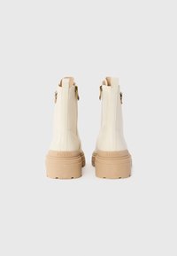Crème-kleurige enkellaarsjes met stevige beige zolen, achterritsen met gouden trekkoorden en "PINKO" branding op de hak.