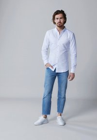 Witte blouse met lange mouwen en kraag, gecombineerd met lichtblauwe versleten jeans en witte sneakers. Eenvoudig, casual ontwerp.