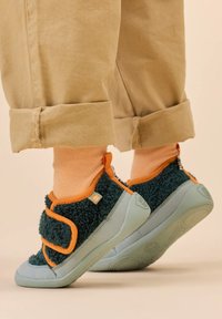 Pantofole morbide di colore verde scuro con suole blu-grigie, accenti arancioni e una strap con Velcro. Indossate con calze rosa chiaro e pantaloni beige arrotolati.