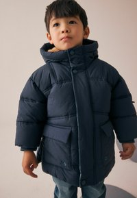 Jongen in een marineblauwe gewatteerde jas met capuchon en voorkruiken, staand tegen een effen lichte achtergrond.