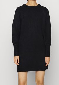 Svart stickad klänning med avslappnad passform, med en ribbad hals och manschetter prydda med små svarta pärlor. Mjuk textur och längd över knäna.