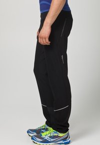 Schwarze Sporthosen aus flexiblem Material mit seitlichen Reißverschlusstaschen und reflektierenden Akzenten, kombiniert mit blauen und grünen Laufschuhen.