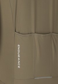 Nahaufnahme eines olivgrünen Stoffes mit drei aufgenähten Taschen und dem vertikal in Weiß gedruckten Wort „ENDURANCE“.