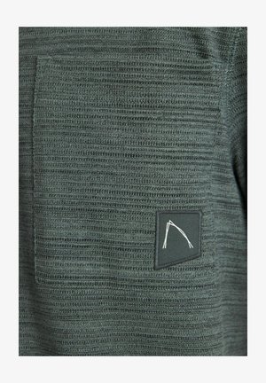 Tume roheline kudumispaita, millel on tekstuuriga kangas horisontaalsete joontega, rinna tasku ja valge mustriga ristkülikuline logo silt.