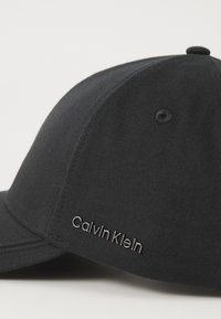 Cappellino da baseball nero realizzato in tessuto liscio. Presenta il logo ricamato "Calvin Klein" in argento sul lato, visiera strutturata e occhio di ventilazione.