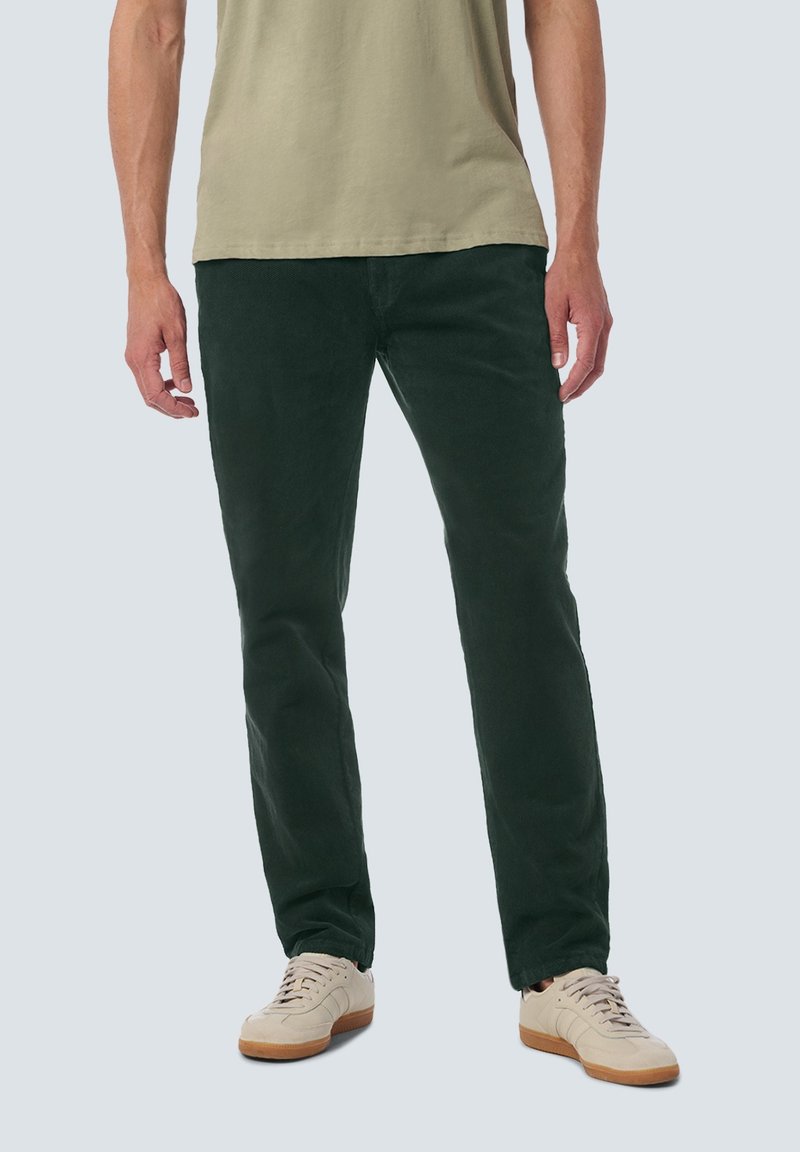 Donkergroene corduroy broek met een recht ontwerp, gecombineerd met lichtgekleurde sneakers, tegen een neutrale achtergrond.