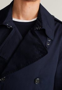 Mężczyzna w granatowym dwurzędowym płaszczu przeciwdeszczowym z zapiętym kołnierzem i ciemnym swetrze z białym wykończeniem przy szyi pod spodem.