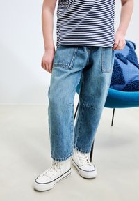 Jasnoniebieskie dżinsy o luźnym kroju, z widocznymi kieszeniami i wyblakłą fakturą, zestawione z białymi, wysokimi trampkami z czarnymi akcentami.