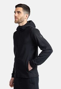 Svart zip-up hoodie med hög krage, tillverkad av slät tyg. Har en diskret logo på vänster axel och sidofickor.