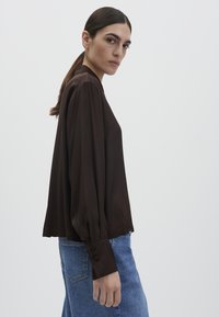 Frau mit dunklem Haar in einem tiefen Pferdeschwanz, die eine lockere dunkelbraune Bluse mit geknöpften Manschetten und blaue Jeans trägt, seitlich stehend vor weißem Hintergrund.