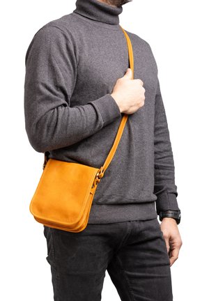 Man draagt een grijze coltrui en zwarte broek, met een kleine mosterdgele leren schoudertas over de schouder.