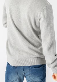 Pull gris clair avec un motif en maille texturée, poignets et ourlet côtelés ; porté avec un jean bleu, affichant une coupe décontractée et un style casual.