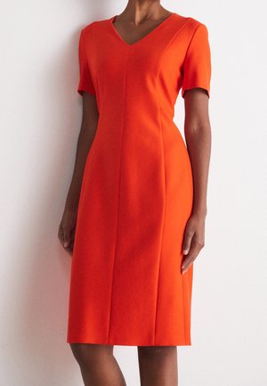 Kvinde iført en knælængde, tætsiddende, lys orange kjole med korte ærmer og V-hals, stående mod en hvid baggrund.