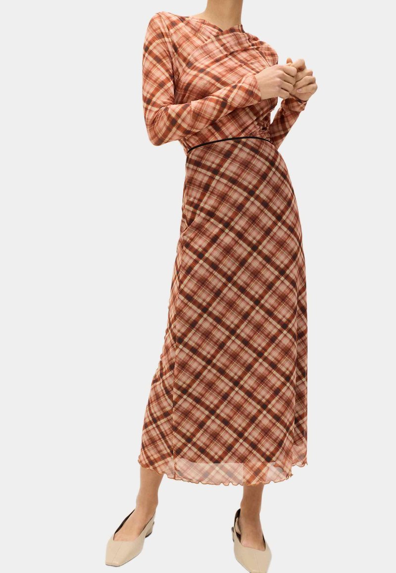 Vrouw draagt een lang, mouwloos jurkje met een rood- en bruin geruit patroon en beige schoenen met lage hak.