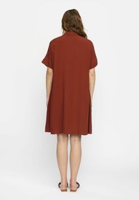 Rostfarbendes Kurzarmkleid mit lockerer Passform, Kragen und seitlichen Schlitzen. Aus glattem Stoff, der bis zur Mitte des Oberschenkels reicht.