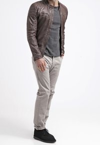 Uomo in piedi con le mani nelle tasche, che indossa una giacca di pelle marrone, una maglietta grigia, pantaloni beige e scarpe nere, su uno sfondo bianco.