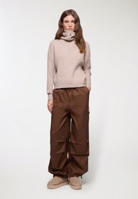 Maglione a collo alto beige a costine con dettagli con zip, abbinato a pantaloni cargo larghi marroni e scarpe slip-on beige.