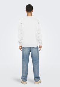 Felpa bianca con maniche lunghe, scollo rotondo e polsini a coste, abbinata a jeans di denim azzurro chiaro, larghi, e scarpe beige.