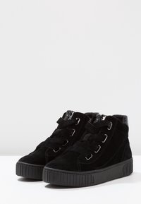 Pair of black high-top sneakers with thick rubber soles and wide velvet laces, displayed on a white surface with gray background.