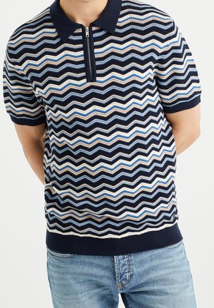 Mann in einem kurzärmligen dunkelblauen Polohemd mit bunten Zickzackmustern und hellblauen Jeans, die Hände hinter dem Rücken.