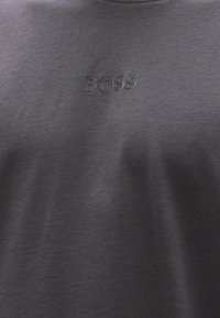 Camiseta de cuello redondo color gris oscuro hecha de tela suave, con un logotipo negro "BOSS" en relieve en el pecho. Textura lisa, diseño informal.