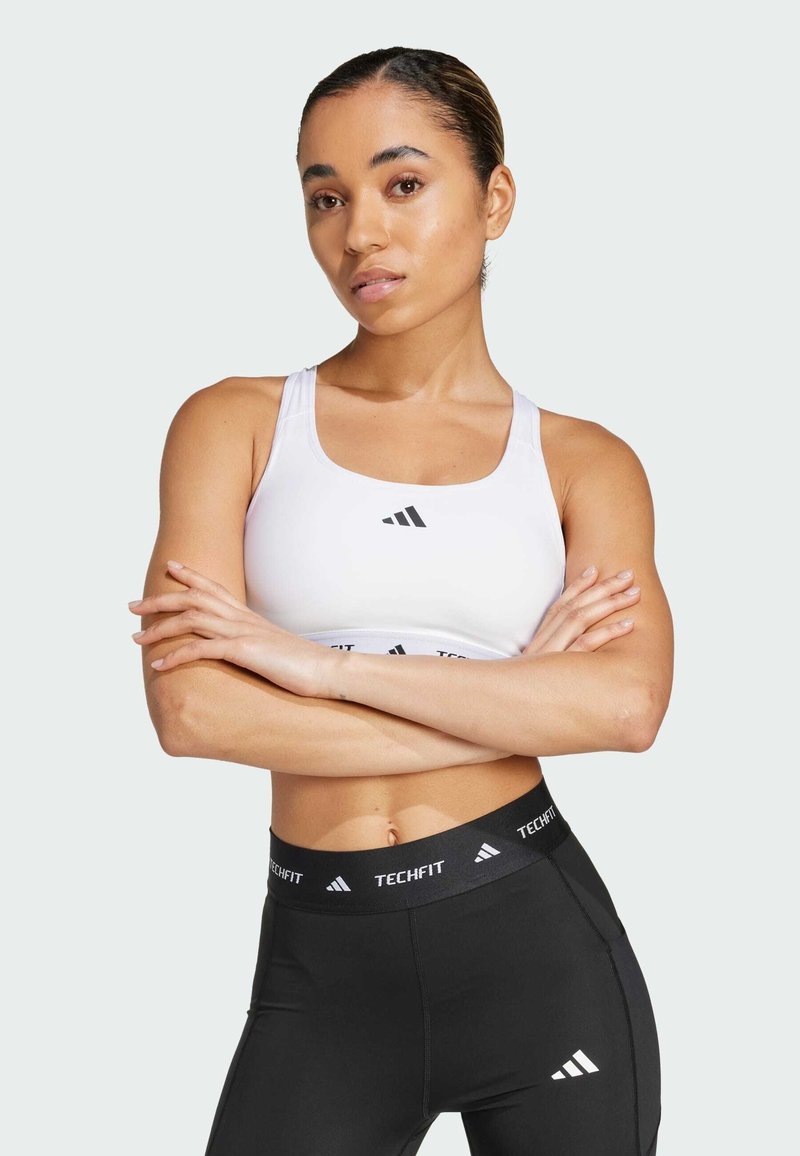 Hvit sports-BH med svart logo, kombinert med svarte leggings med en merkevarebelte. Glatt tekstur, tettsittende design for treningstøy.